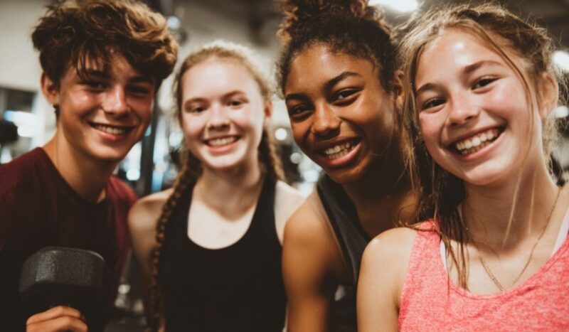 Group of smiling teenagers at a gym holding a dumbbell and posing together