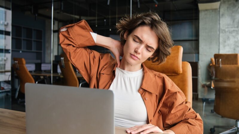Woman at a desk rubs her neck while working on a laptop