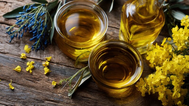 Olive oil and canola oil sit in glass containers on a wooden table with fresh herbs and flowers