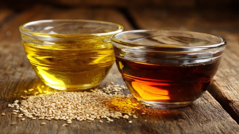 Olive oil and sesame oil sit in glass bowls beside sesame seeds on a wooden surface