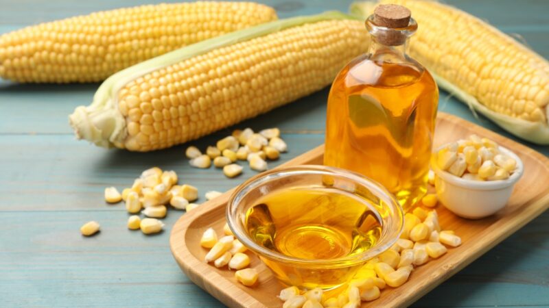 Corn oil sits beside fresh corn and loose kernels on a wooden tray