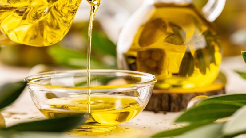 Extra virgin olive oil flows into a glass bowl beside fresh olive leaves