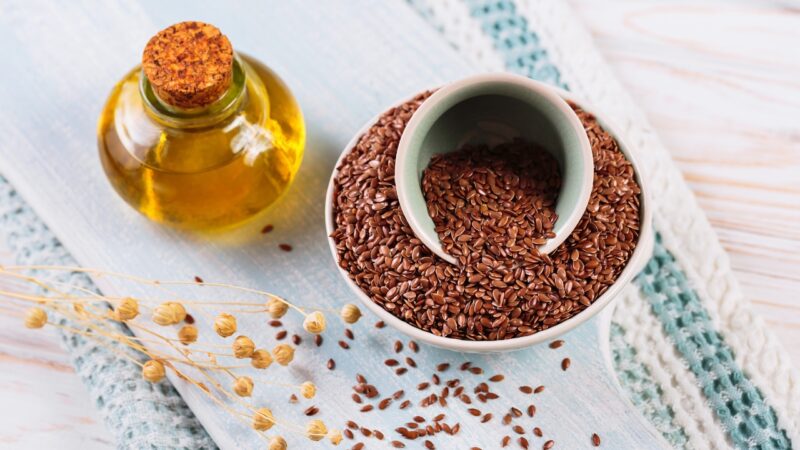 Flaxseed oil sits beside a bowl of whole flaxseeds on a light wooden surface