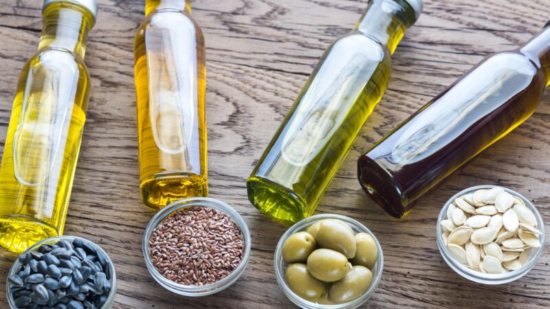 Several vegetable oils are shown in glass bottles beside olives, flaxseeds, and sunflower seeds