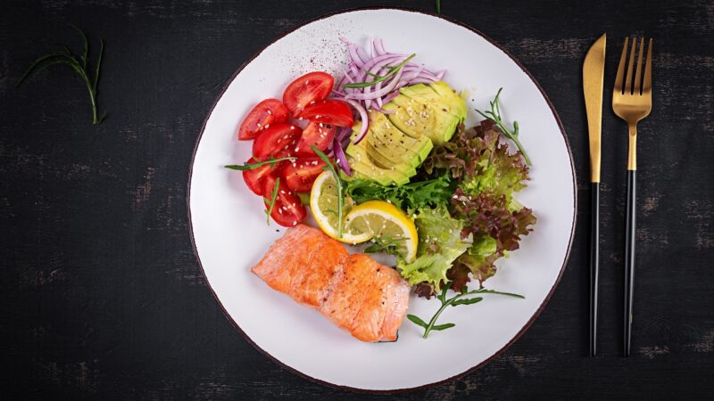 Paleo salmon meal with avocado, tomatoes, greens, and lemon on a dark table