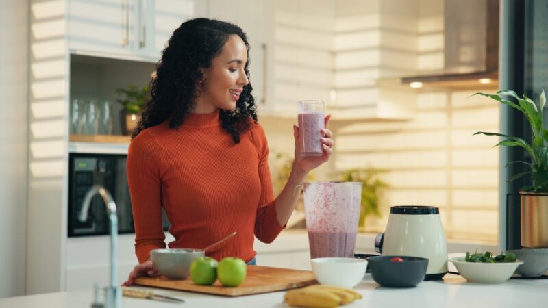 Woman holds a smoothie beside a blender in a bright kitchen