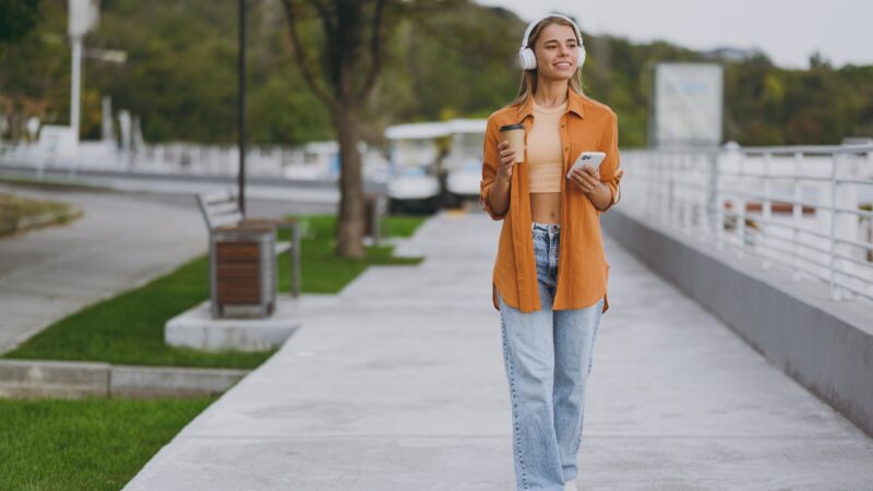 Woman with headphones walks outdoors with a drink and phone