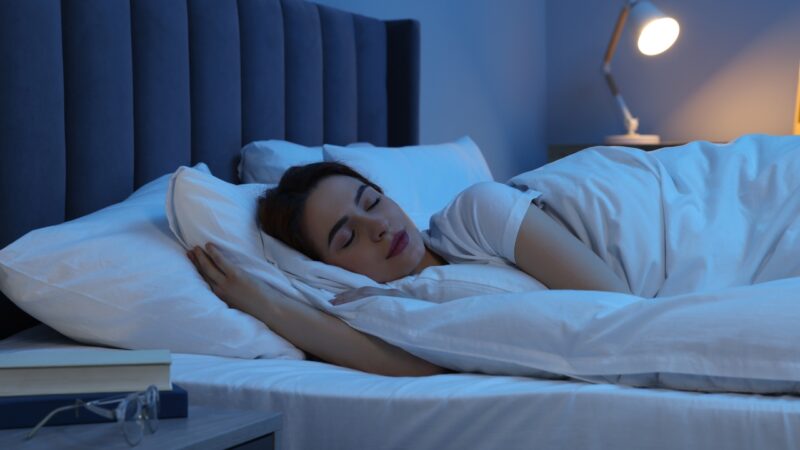 A woman sleeps in bed at night with a bedside lamp on