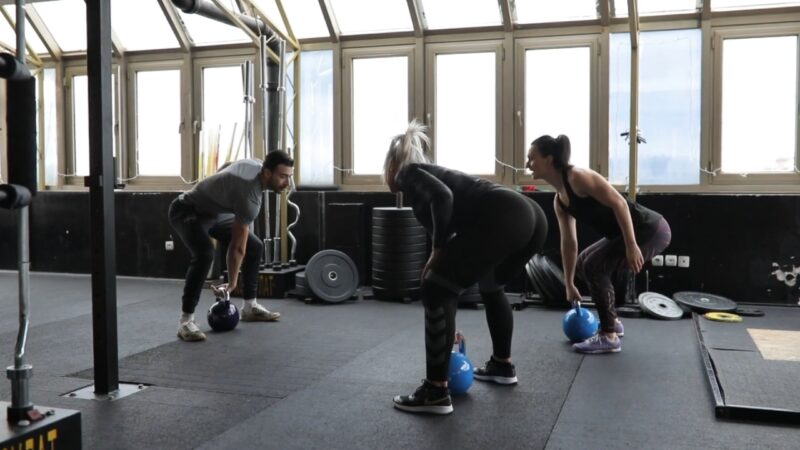 A fitness trainer guides two clients through kettlebell exercises at the gym