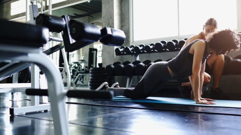 A personal trainer supports a tired client through a floor exercise at the gym